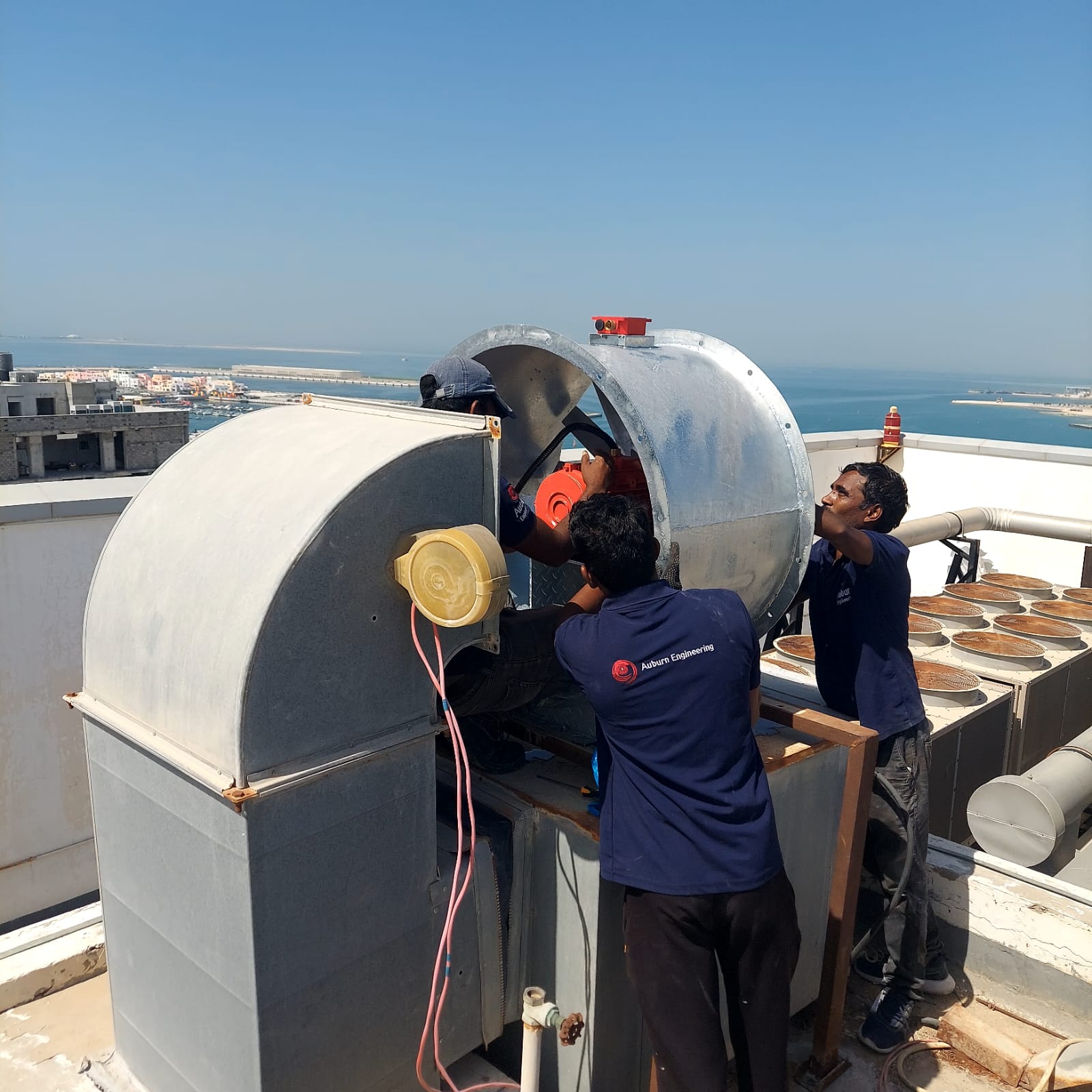 Two Auburn technicians installing a rooftop ventilation fan above Doha's coastline